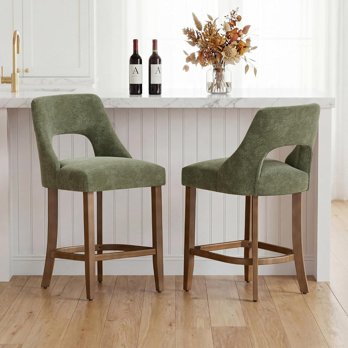 Two green upholstered counter stools with wood legs side-by-side at a white island topped with bottles and a vase.