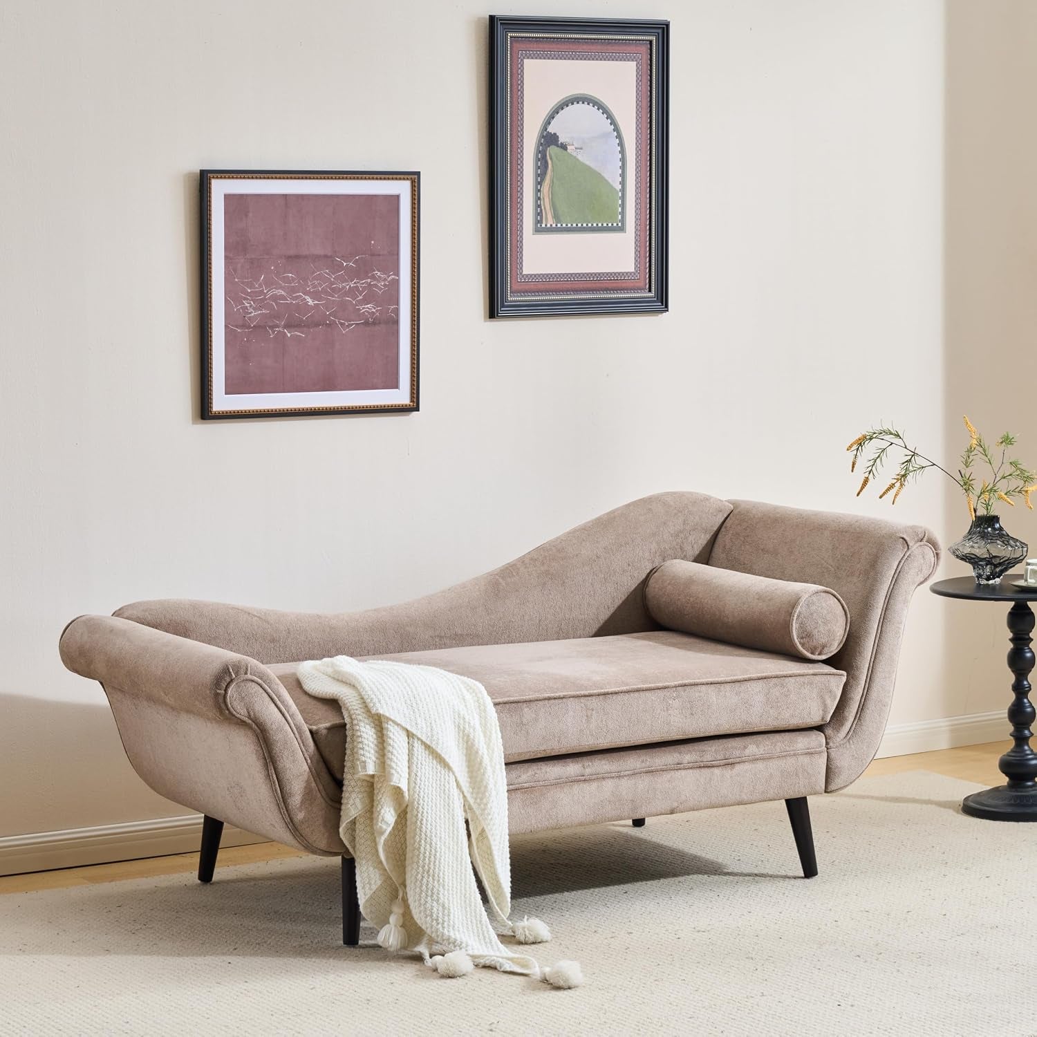 Angled side view of the light brown chaise with bolster pillow and throw beside a small round side table.