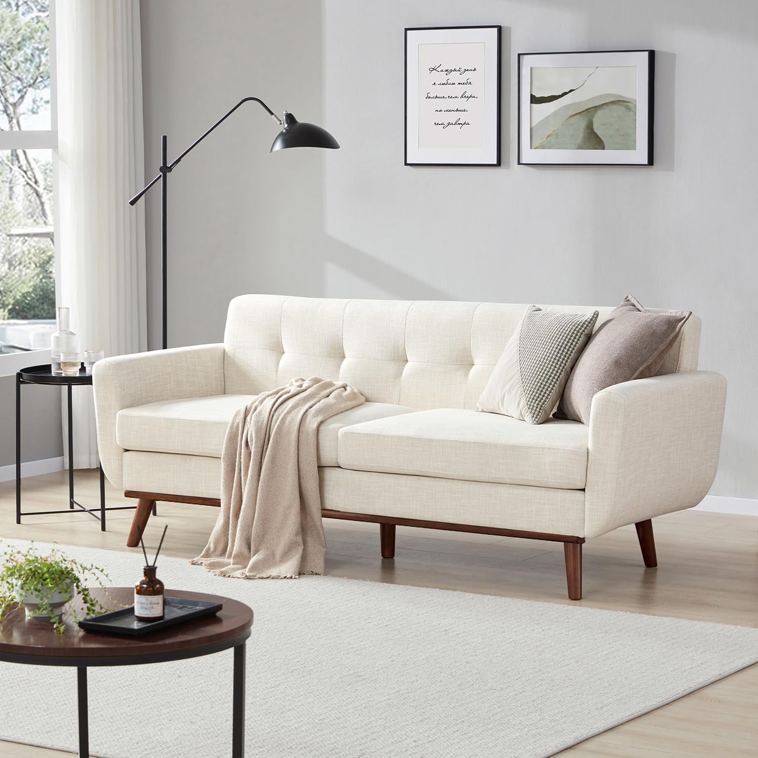 Beige button-tufted loveseat in a bright living room with wooden legs and throw blanket.