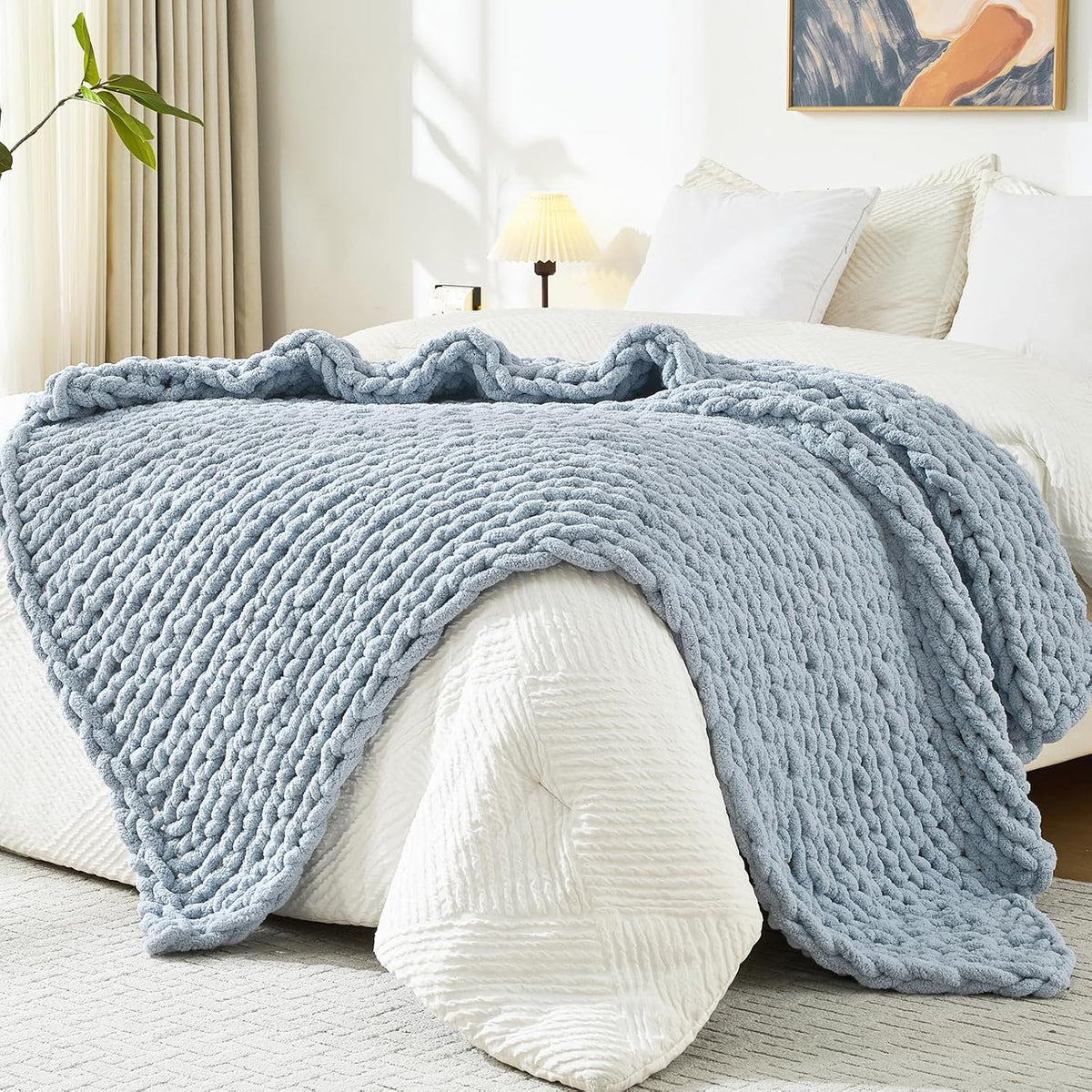 Pale blue chunky knit throw draped over a white bed with pillows and a bedside lamp