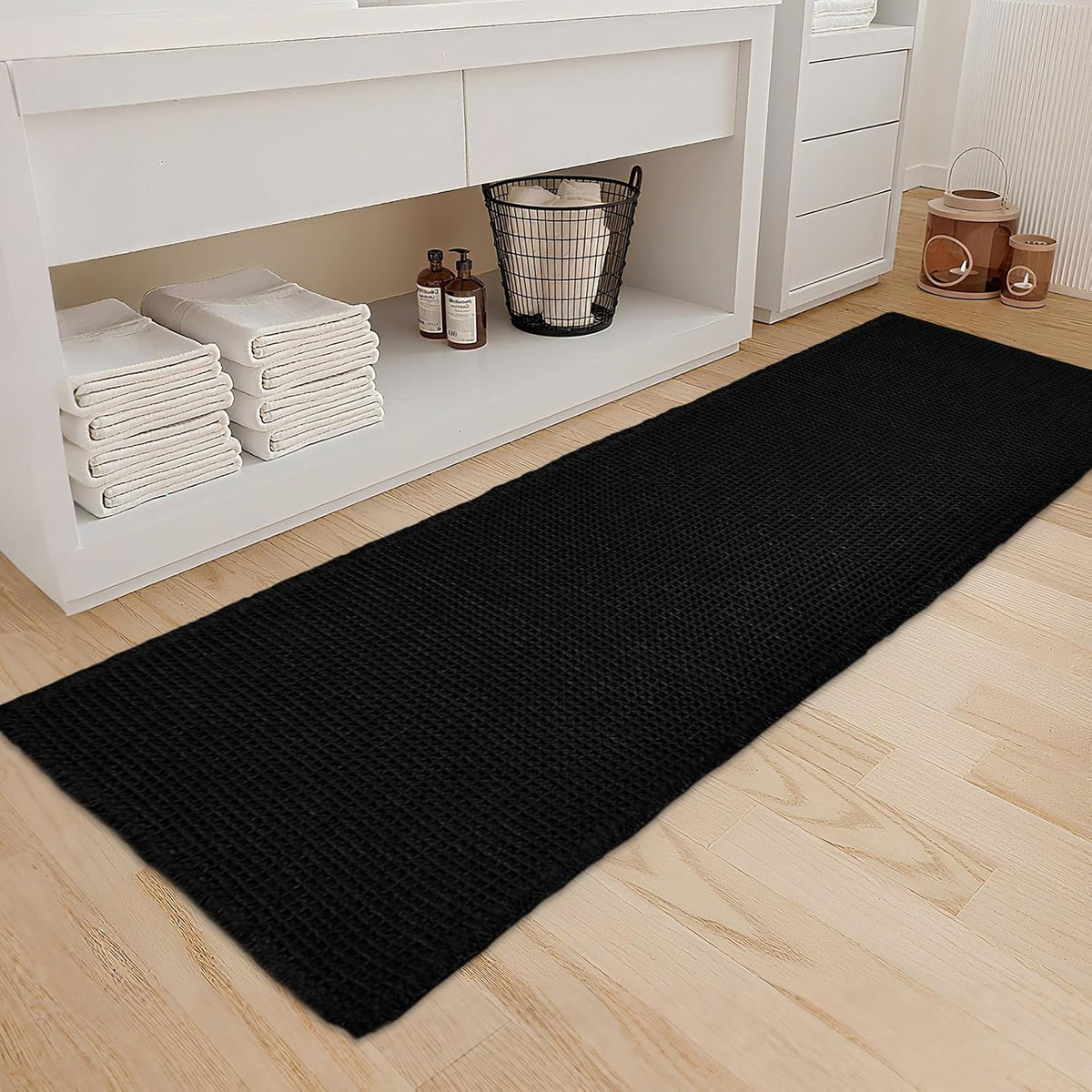 Black waffle runner-style bath mat laid on a light wood floor next to white shelving with towels.