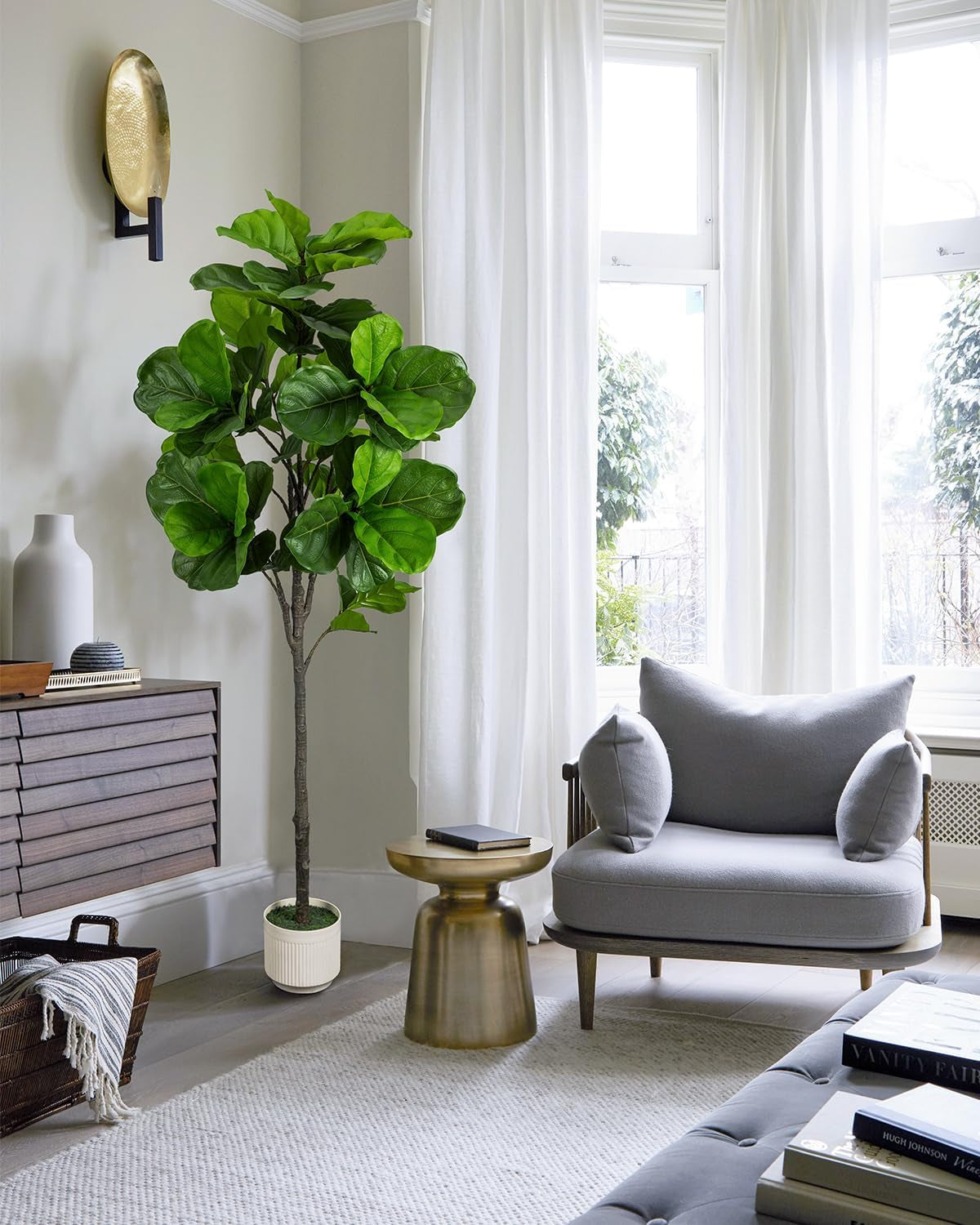 Six-foot artificial fiddle leaf fig in a ribbed white planter beside a wooden sideboard and gray lounge chair by tall windows