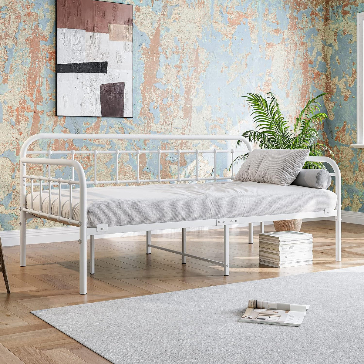 White metal daybed dressed in light bedding set against a distressed blue-and-peach accent wall with a potted plant nearby.