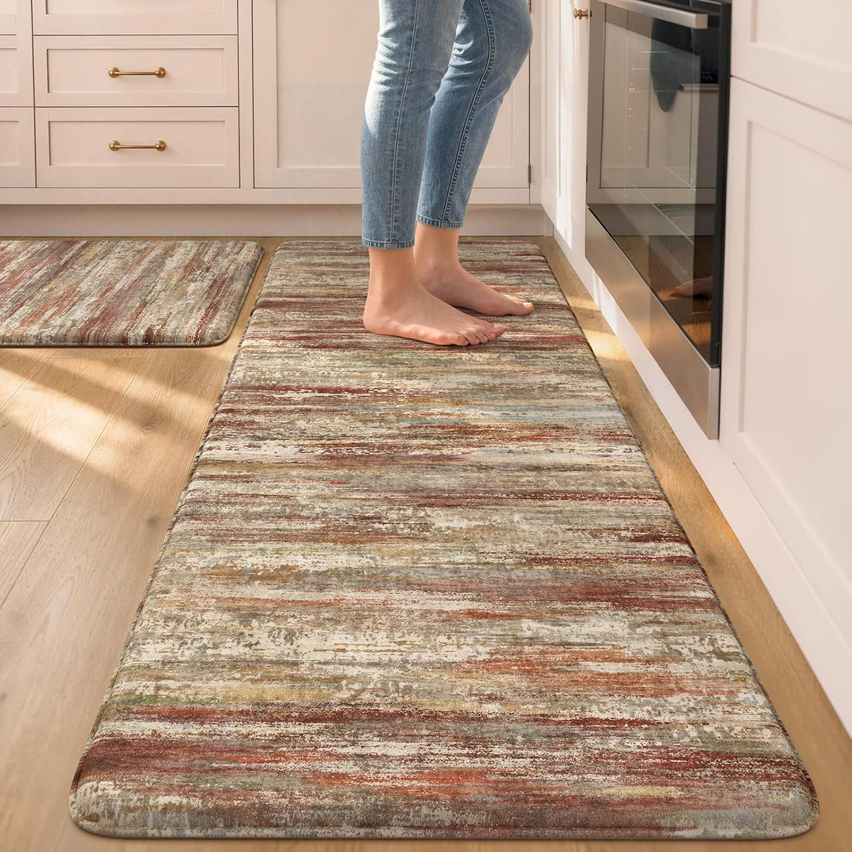 Person standing barefoot on a long multicolored striped runner mat next to cabinets and an oven; a smaller mat sits behind.