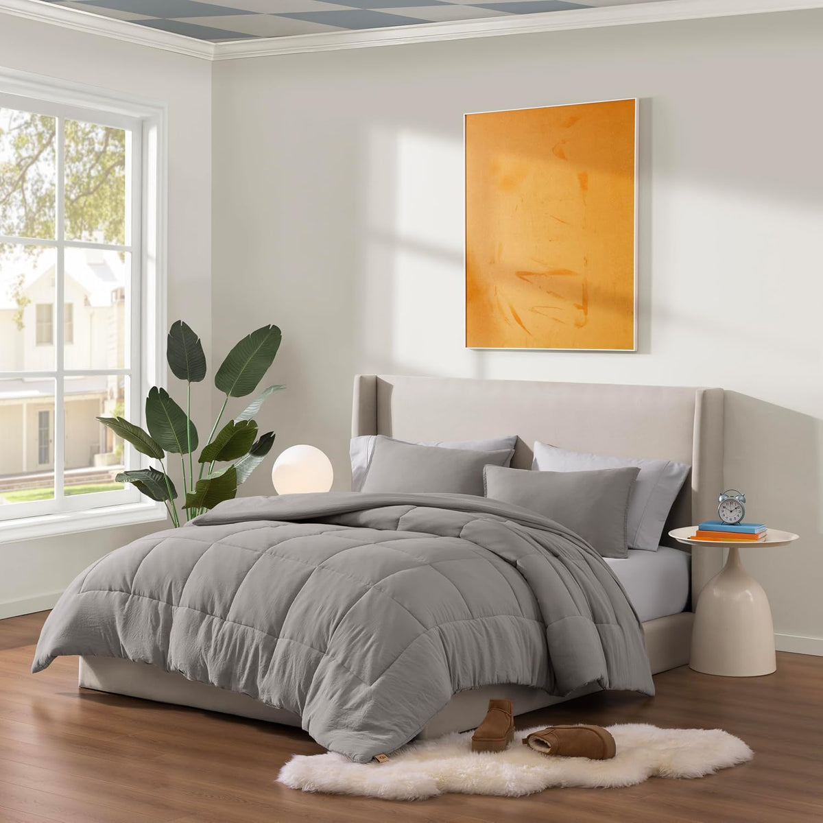 Bedroom scene with light grey box-stitched comforter draped over bed and slippers on floor