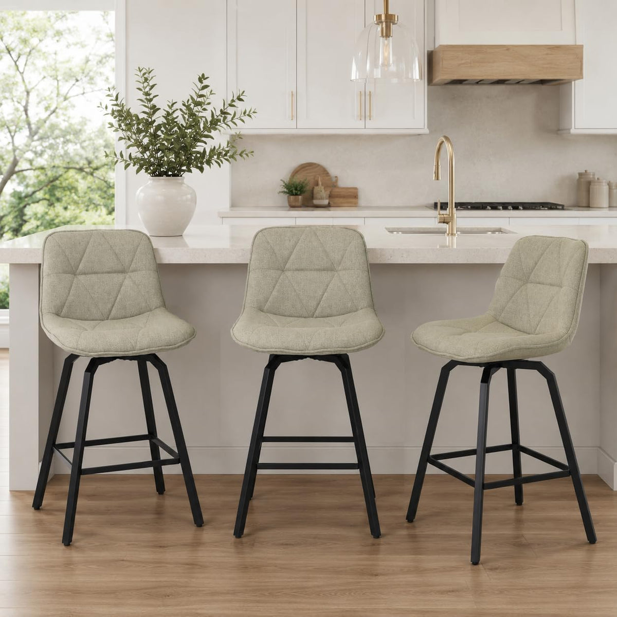Three light beige fabric-upholstered counter stools with black metal legs placed along a kitchen island in a bright kitchen.