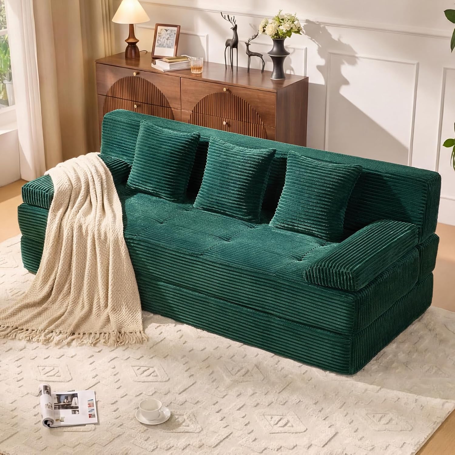 Front three-quarter view of a dark green corduroy three-seat sofa with three square back pillows and a cream throw.