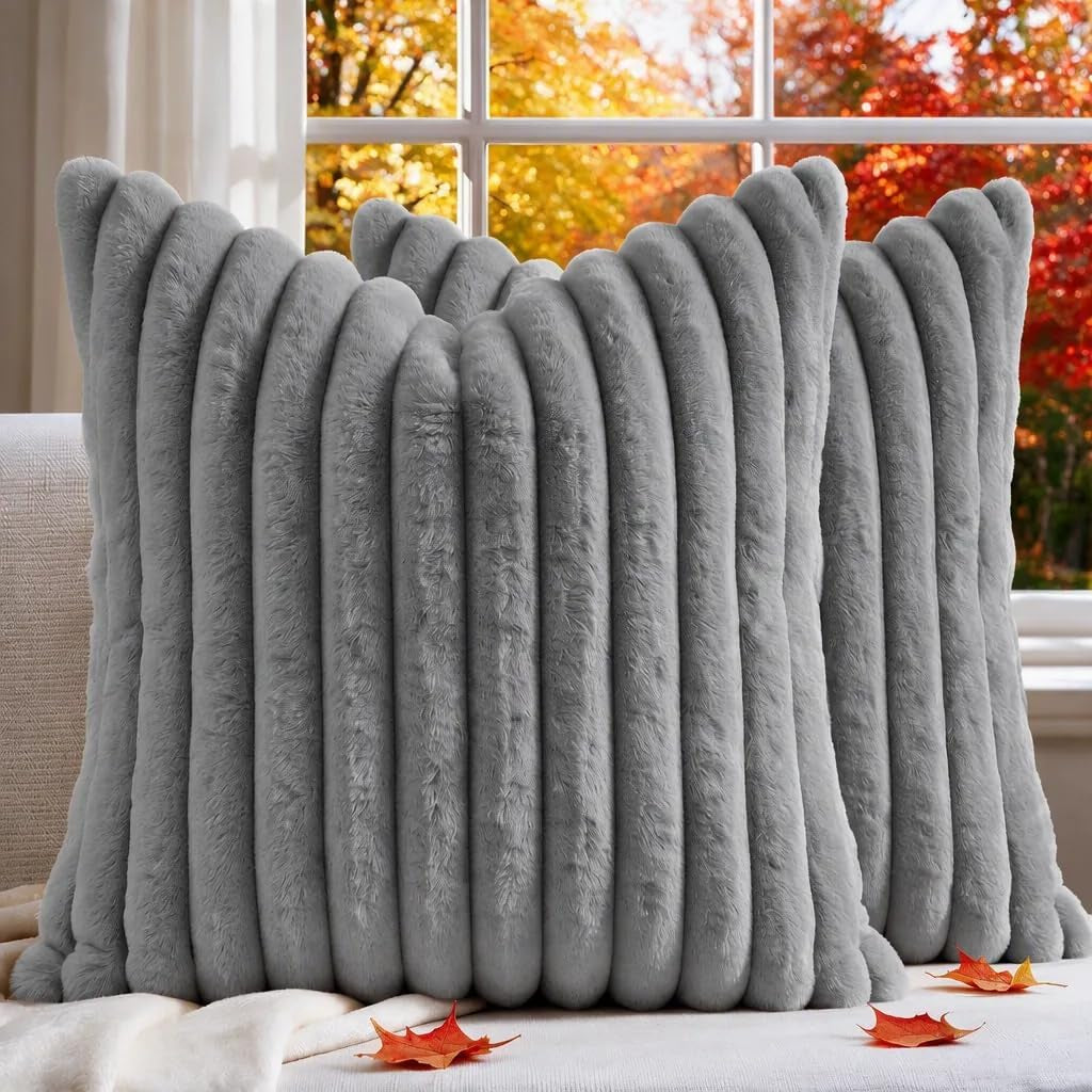 Pair of ribbed faux-fur square pillows in light gray on a sofa with autumn leaves and window behind.
