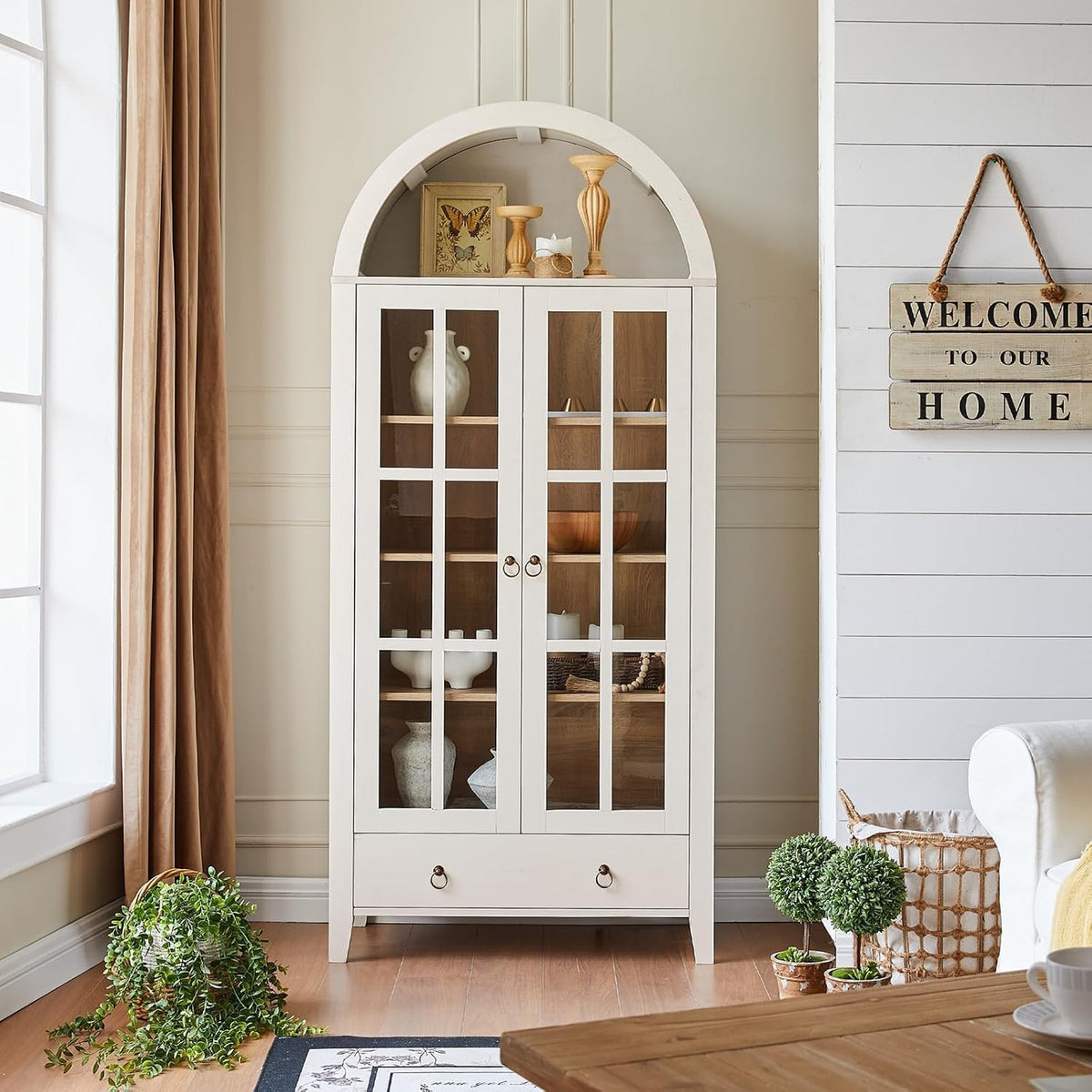 White arched cabinet with glass doors and bottom drawer placed against a neutral wall with plants.
