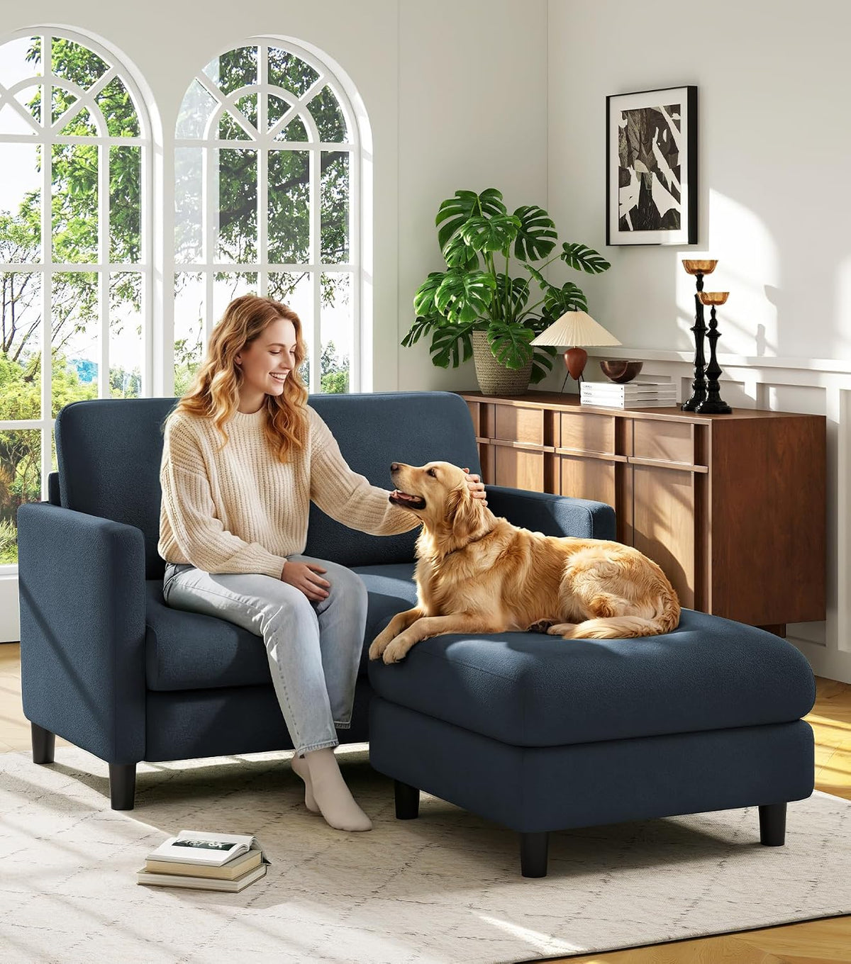 Blue-grey loveseat with matching ottoman in a sunlit room; woman pets a golden retriever lying on the ottoman.