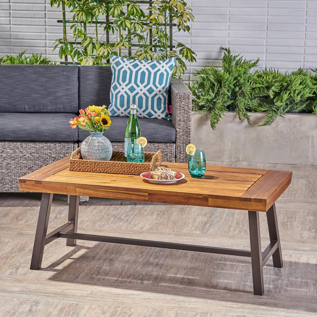 Acacia slat-top coffee table in patio setting with tray, glassware and rattan sofa in background