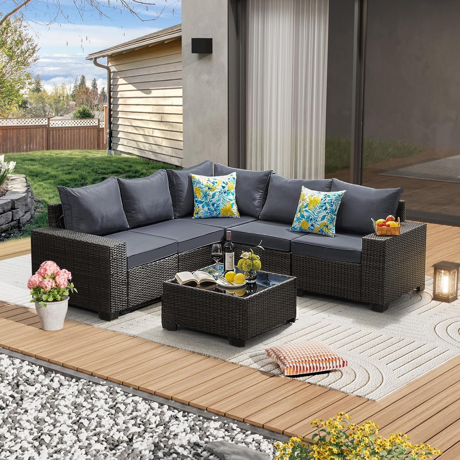 Outdoor L-shaped rattan sectional with dark gray cushions and glass-top coffee table on a deck.