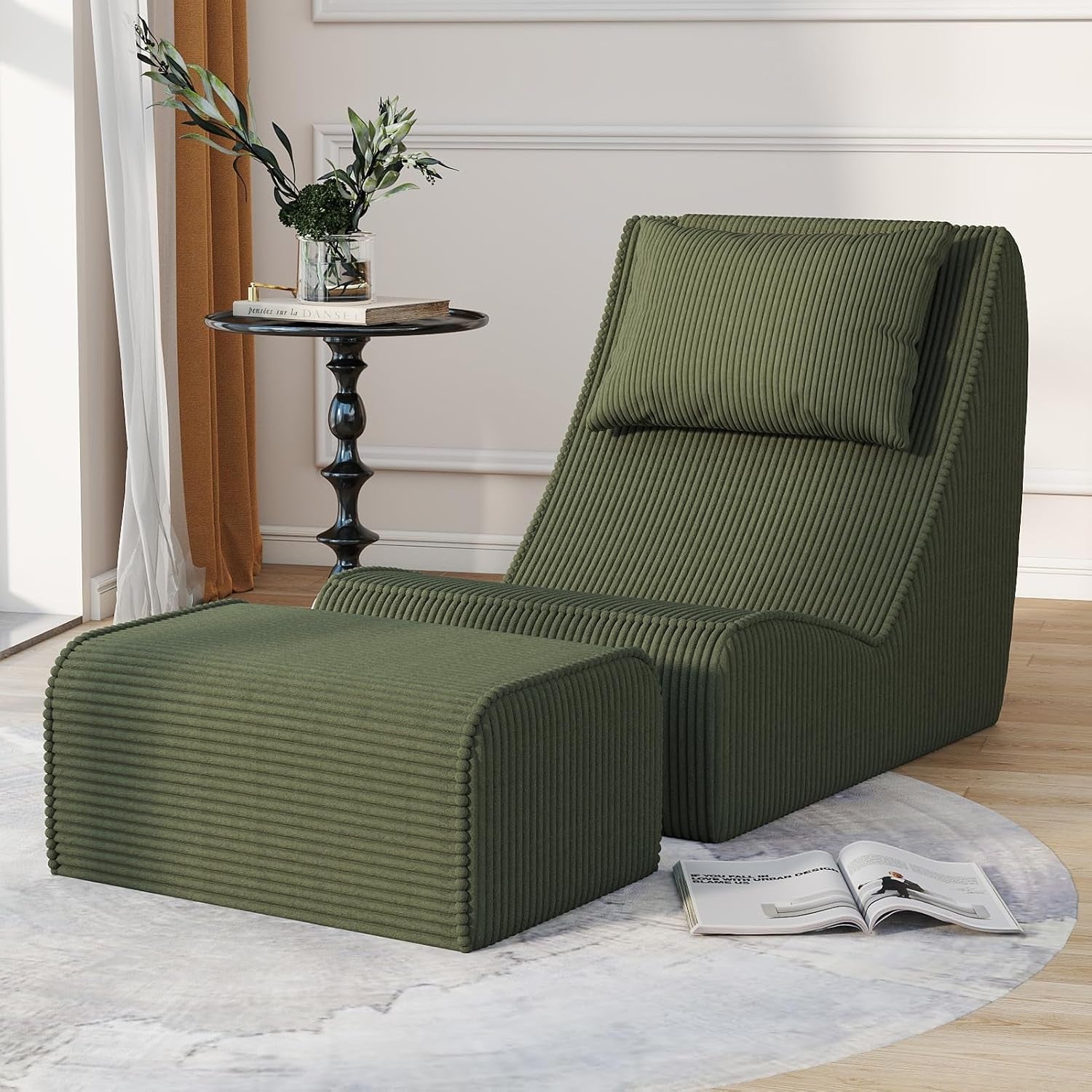 Olive green chaise lounge with ottoman angled right, an open book on the rug and a slender vase with branches by the window.