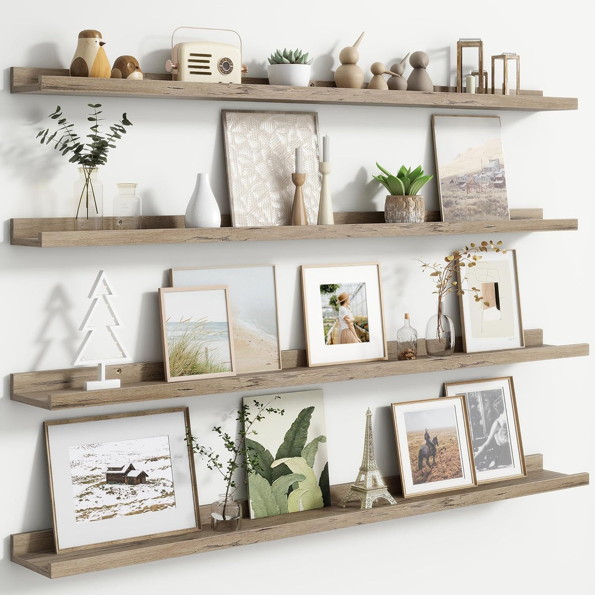 Three long light‑wood picture ledges stacked with framed art, vases and small plants along a white wall.
