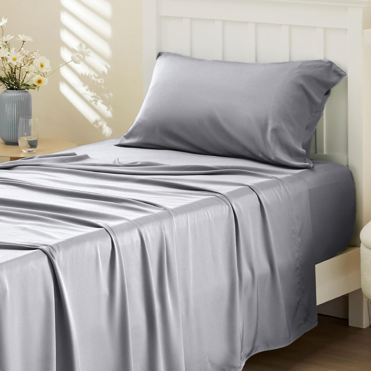 Side view of a single bed with smooth silver-gray sheets and a pillow, sunlight and a vase on a nearby table.