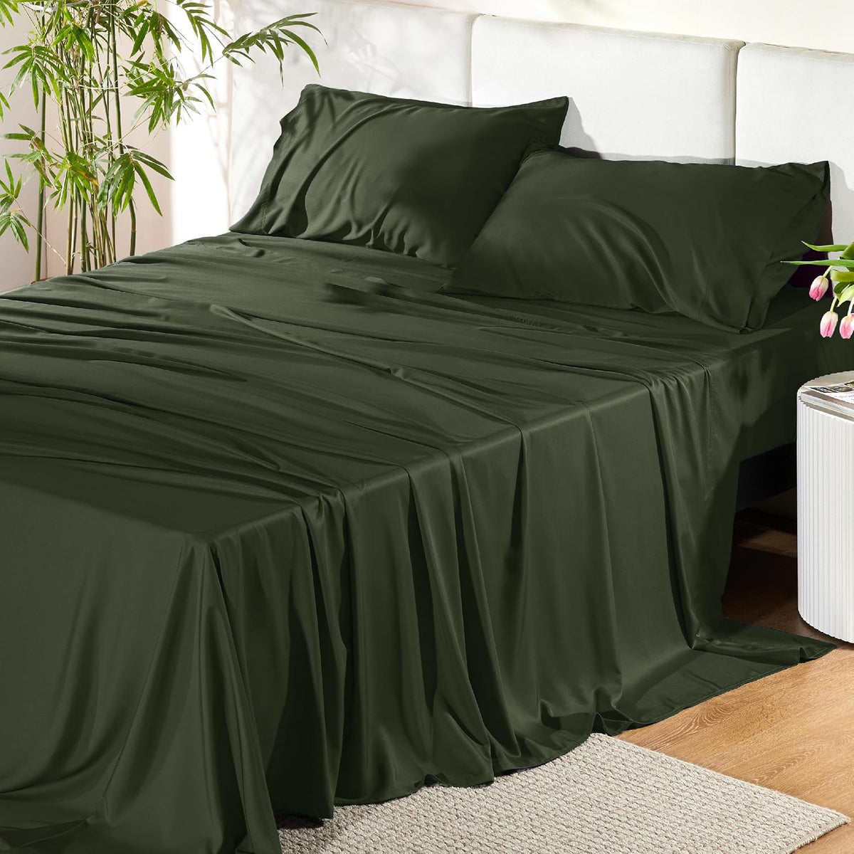 Dark olive-green sheet set draped over a made bed with two matching pillowcases and plants nearby.