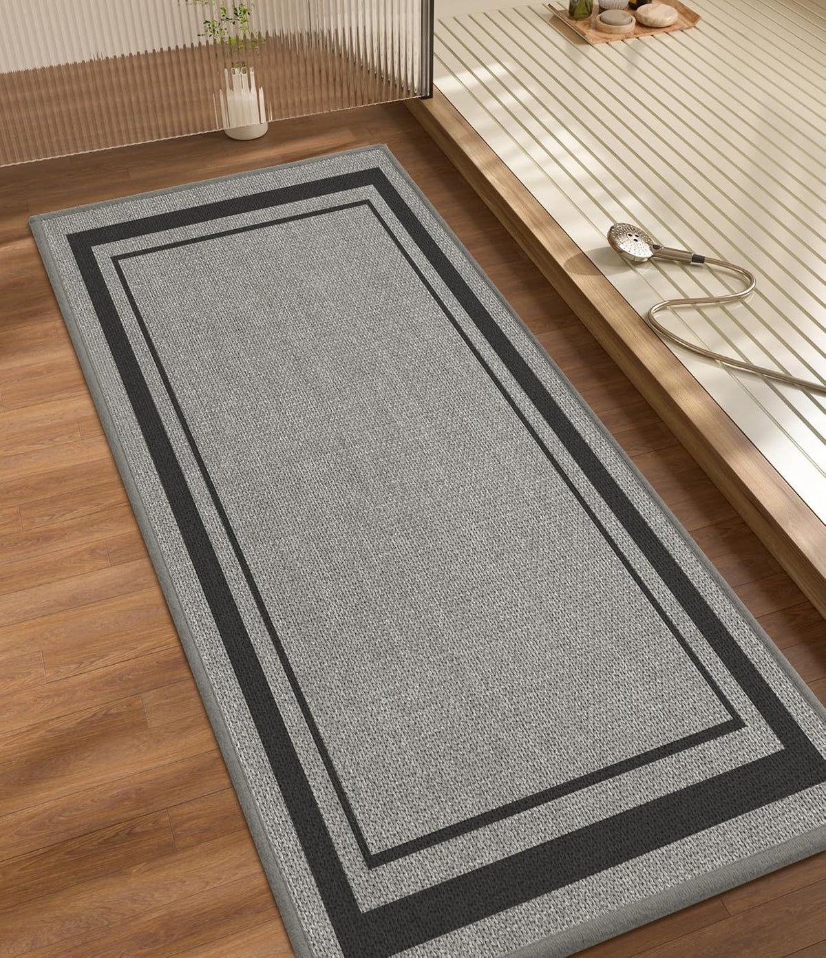 Long rectangular gray bath rug with double border placed on wood floor next to a raised tub area.
