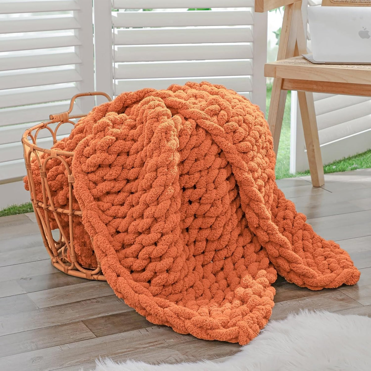 Side view of a burnt-orange chenille chunky knit throw draped from a woven basket onto a wood floor.