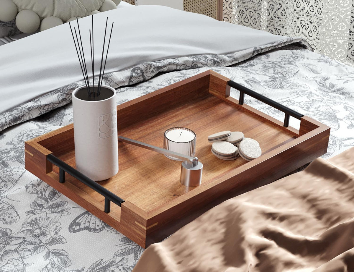 Brown acacia serving tray with black metal handles angled on a bed holding a vase and candle