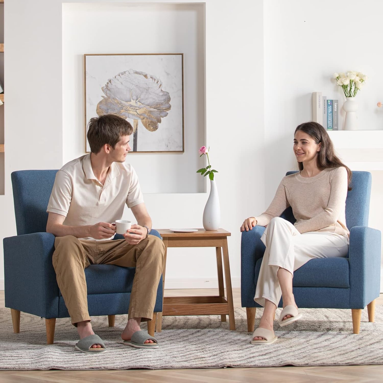 Two matching blue armchairs in a bright room with a wooden side table and two people sitting and talking.