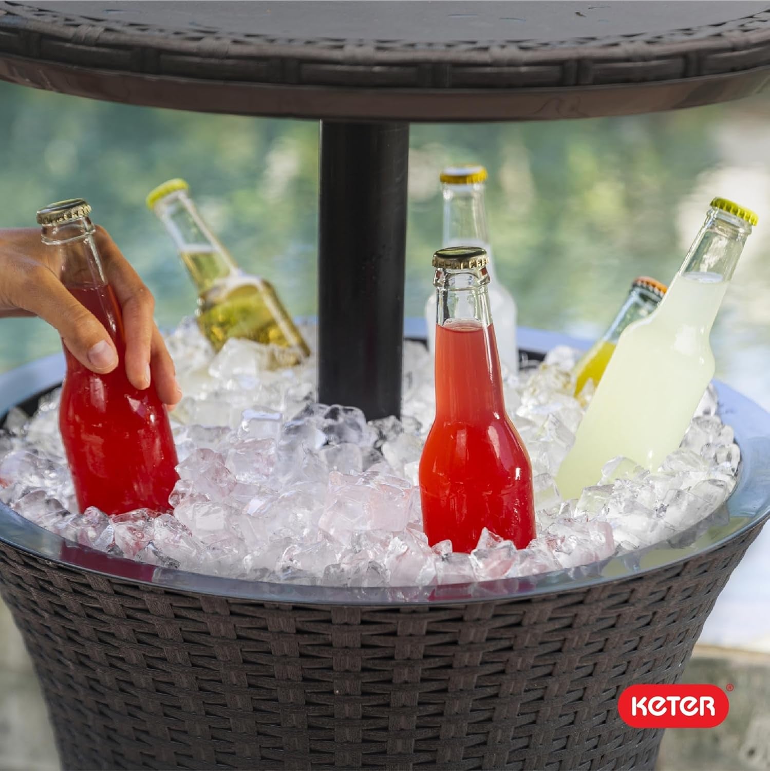 Top view of a round wicker cooler filled with ice and assorted bottled drinks, a hand reaching for a red bottle.