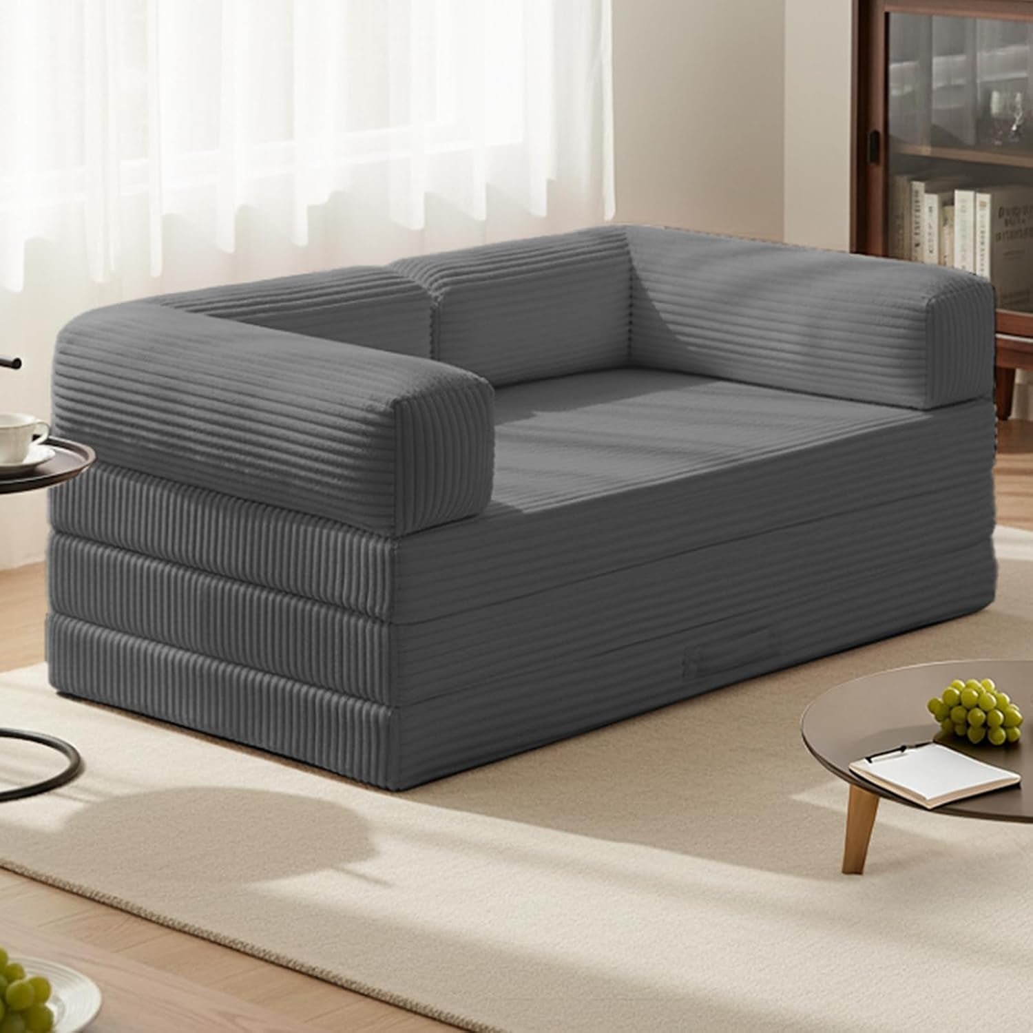 Angled front view of grey tri-fold sofa bed with corduroy cover and L-shaped armrests on a light rug