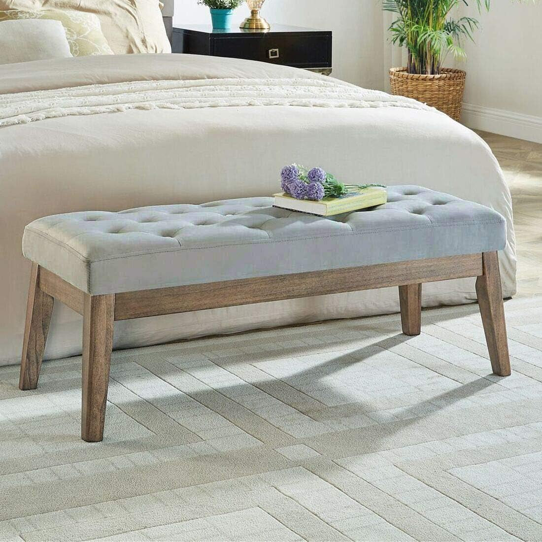 Three-quarter view of a light blue-gray tufted bench with natural wood legs in a bedroom, book and flowers on top.