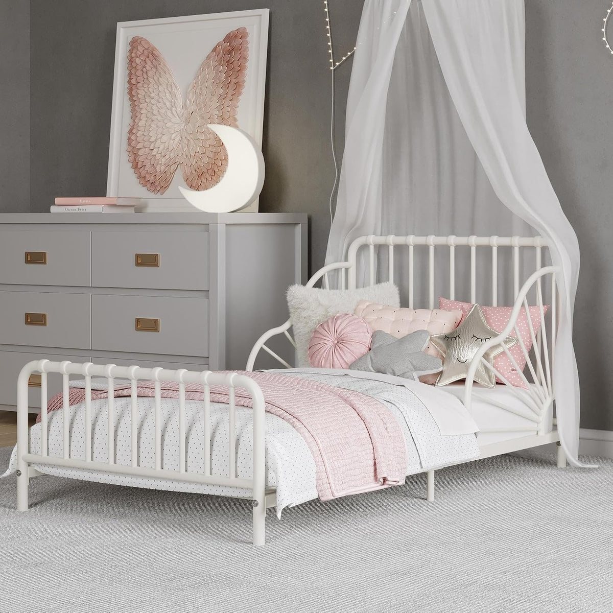 Off-white toddler bed styled in a nursery with pink pillows, canopy and dresser in background.