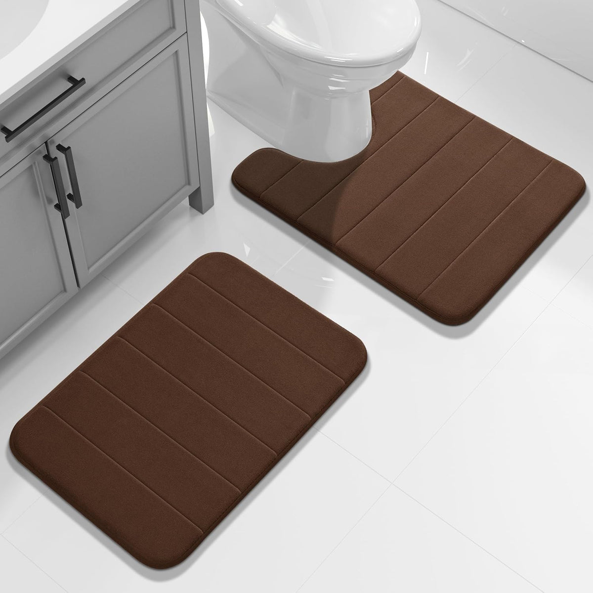Two brown memory-foam bathroom mats on glossy white tile beside a vanity and toilet