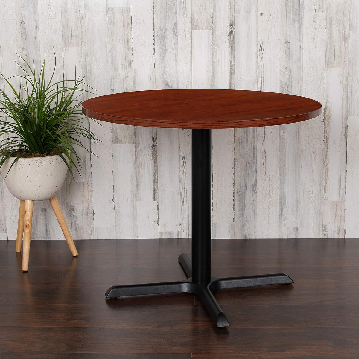 Round cherry laminate table with black X-base photographed against a whitewashed wall, front view