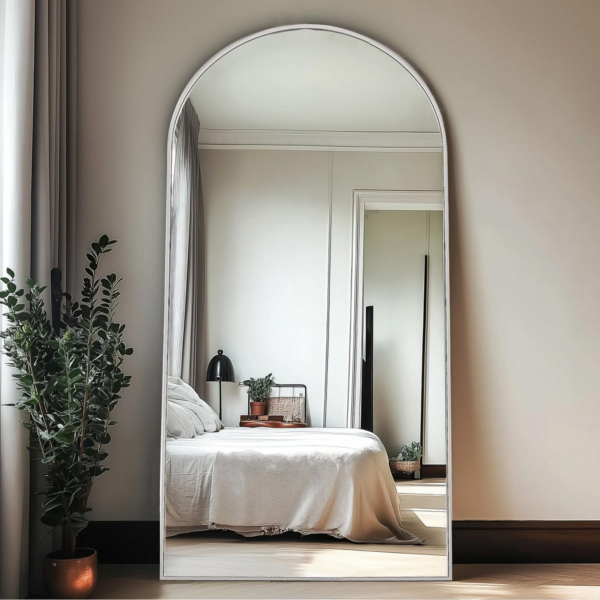 Arched wall mirror with a white frame reflecting a bed and bedside lamp in a neutral bedroom.
