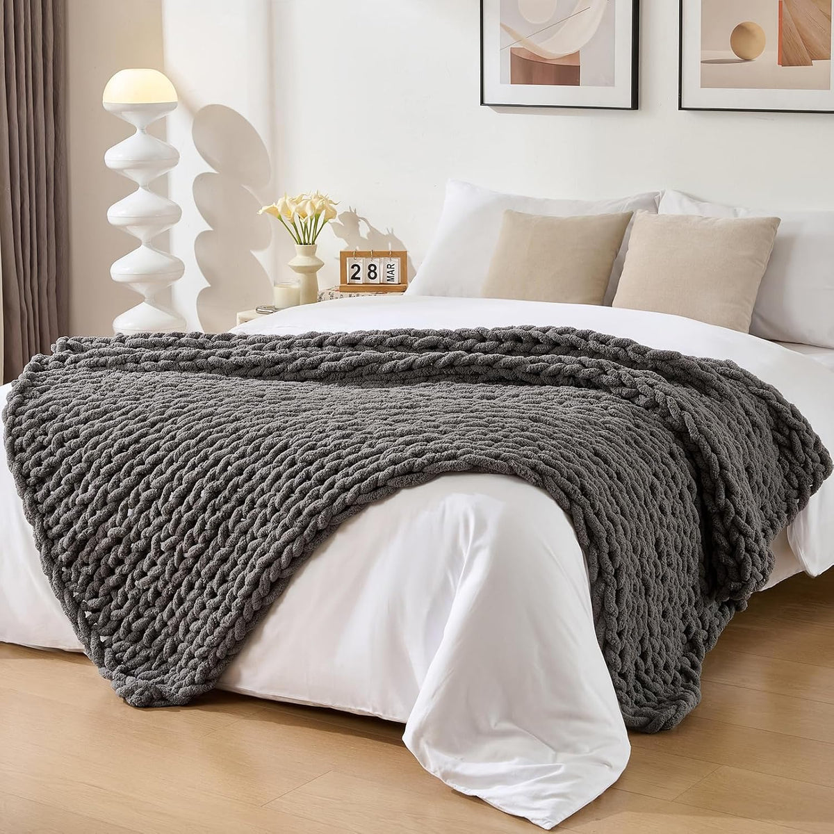 Charcoal-gray chunky knit throw folded across the foot of a made bed with lamp and framed art behind.