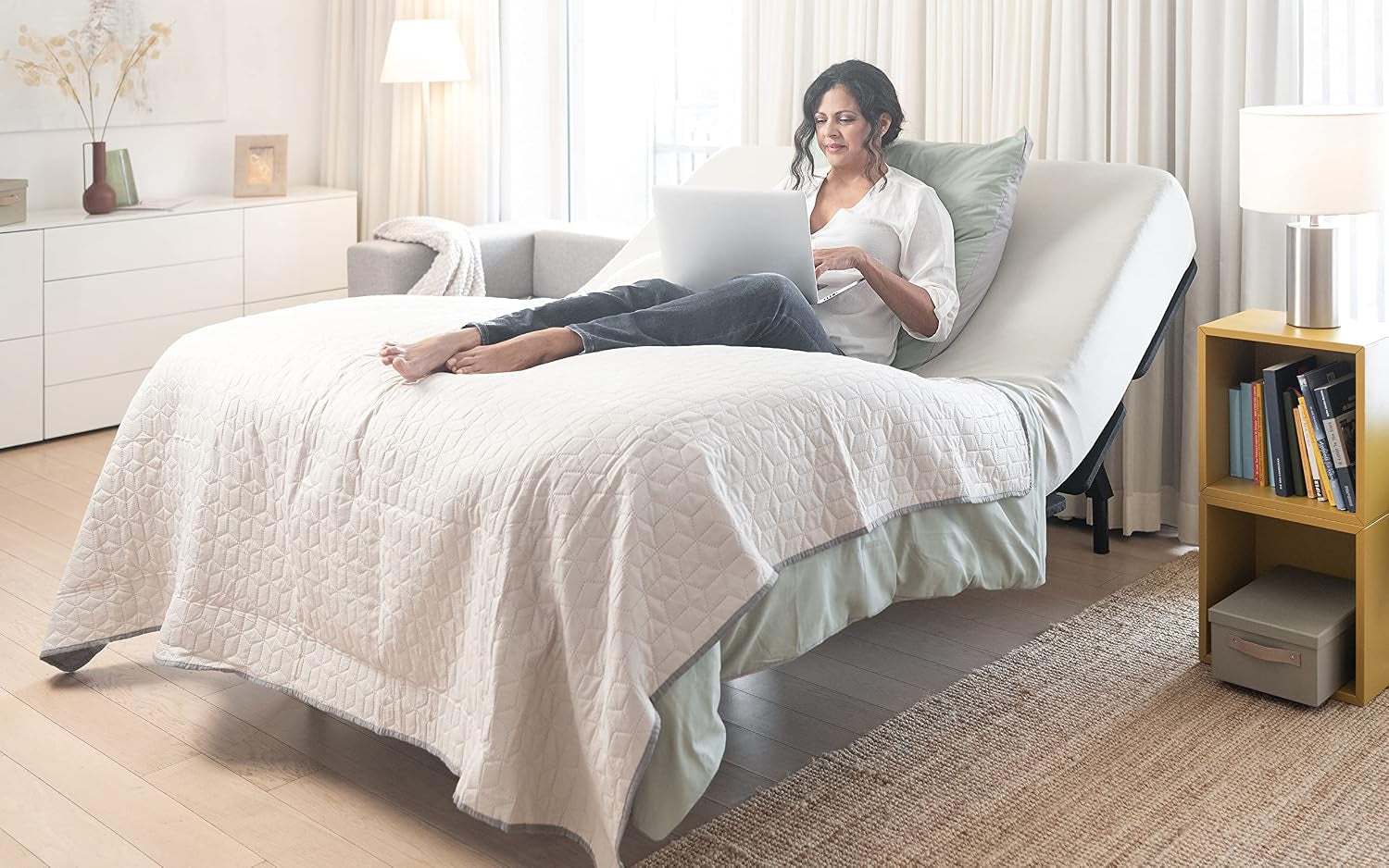 Bedroom scene with woman reclining on raised head section of adjustable bed base while using a laptop.