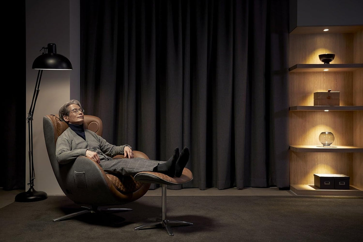 Man reclining in a caramel leather-and-gray fabric massage chair with matching ottoman in a dimly lit living room.