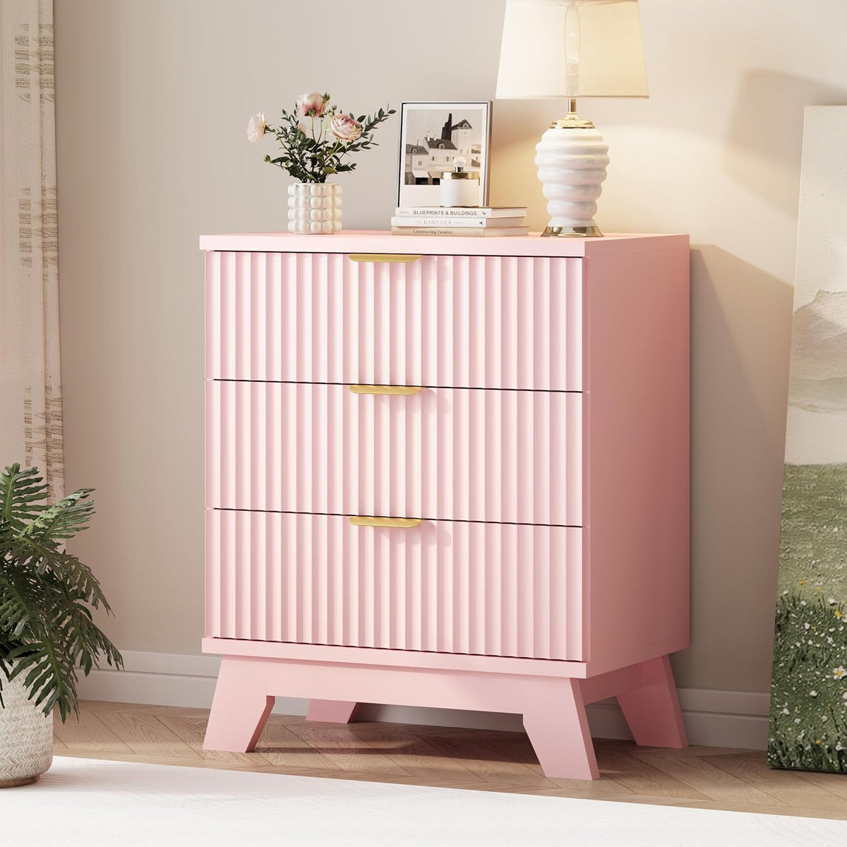 Three-drawer tall chest in pink with fluted drawer fronts and gold handles, styled with lamp and vase.