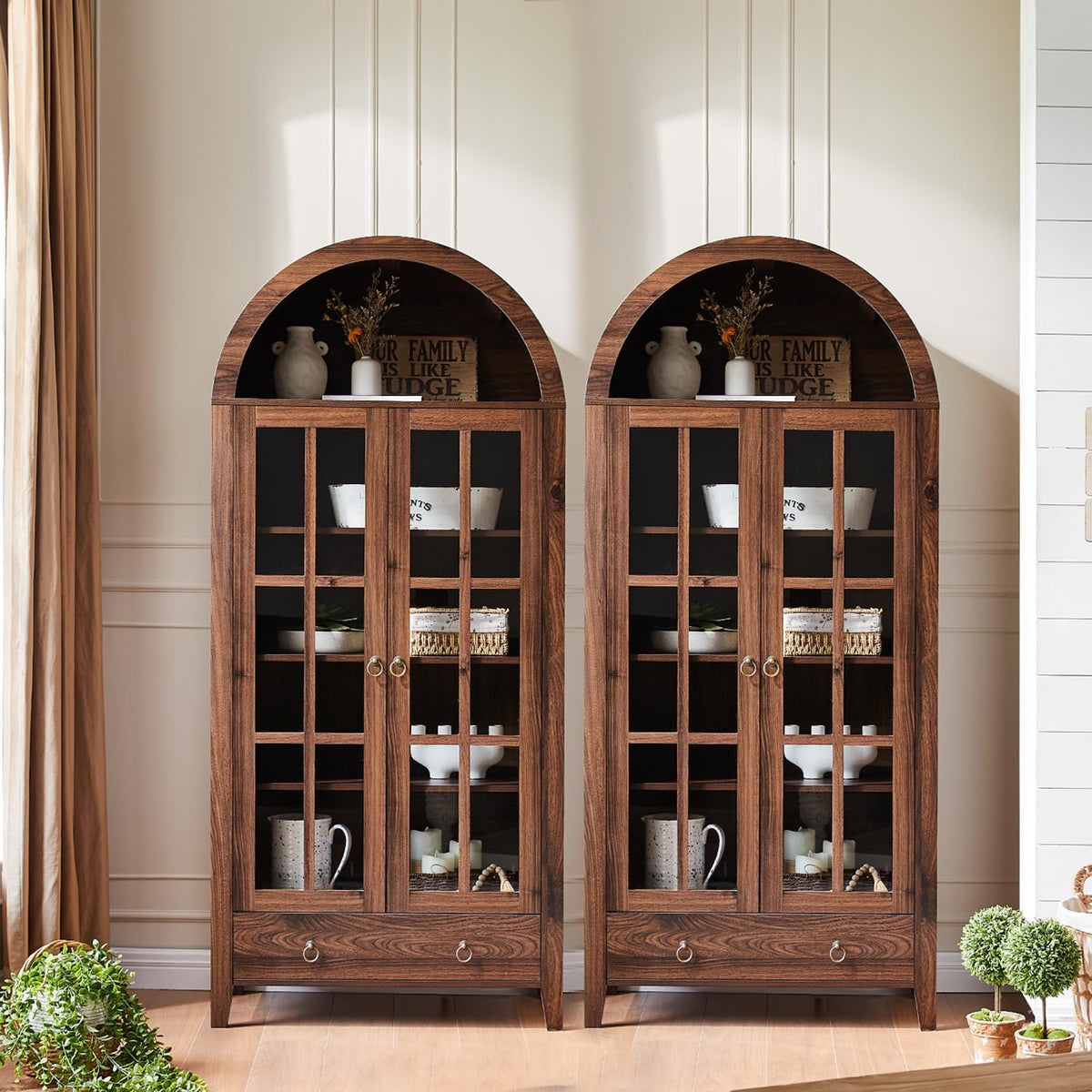 Two arched-top glass-door cabinets placed side-by-side against a wainscoted wall with plants and curtain at the edges
