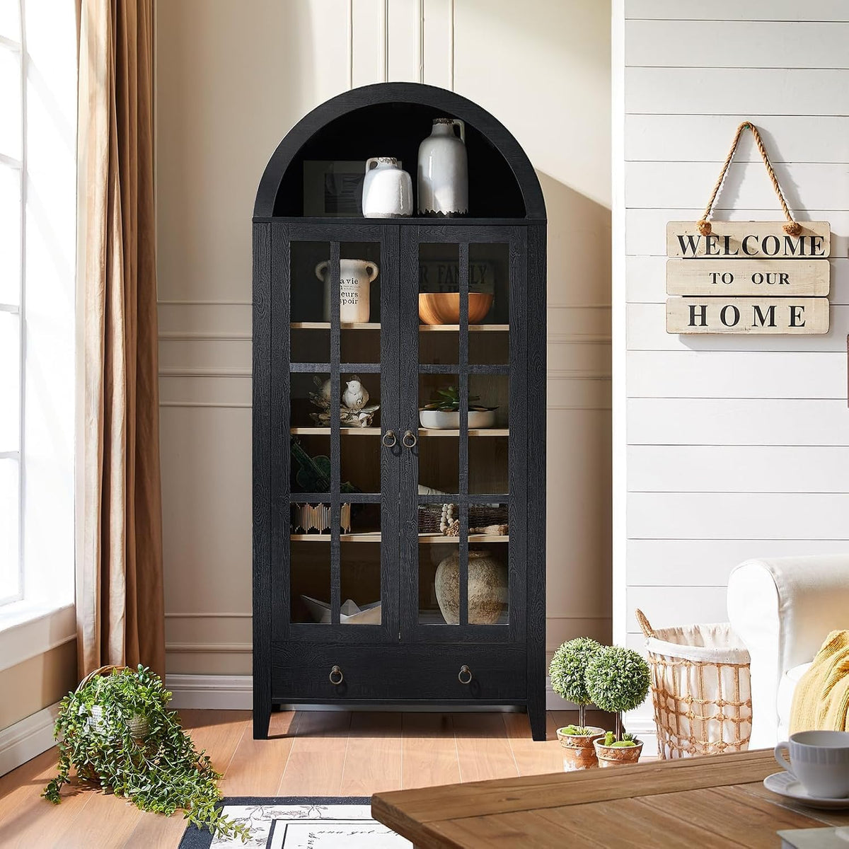 Black arched cabinet with glass-paneled doors and decor, staged between window and shiplap wall.