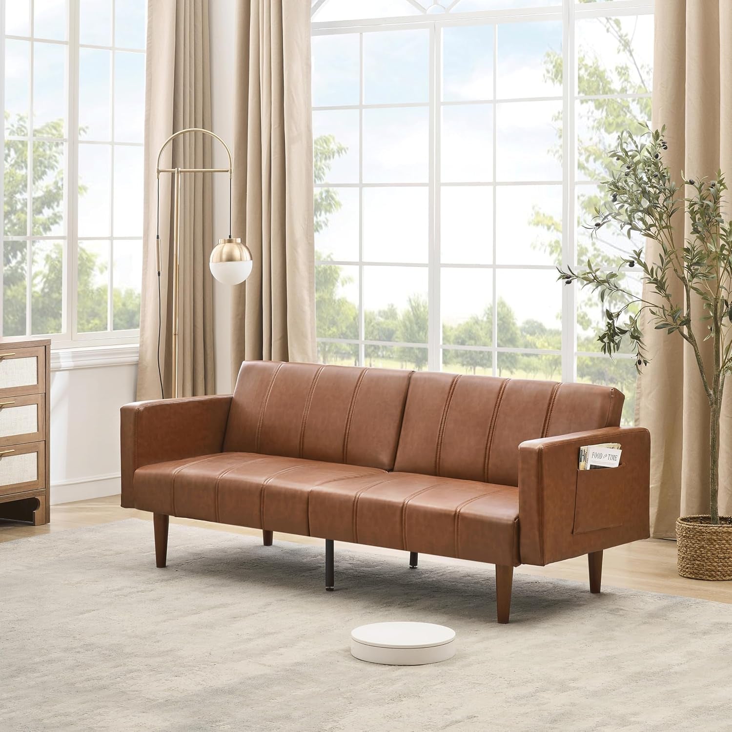Brown PU leather futon sofa in a bright living room with tall windows and a floor lamp