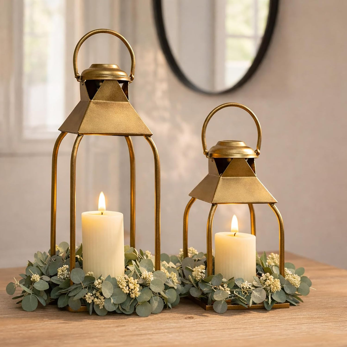 Close view of two antique gold lanterns with lit pillar candles and eucalyptus wreath on a wooden table.