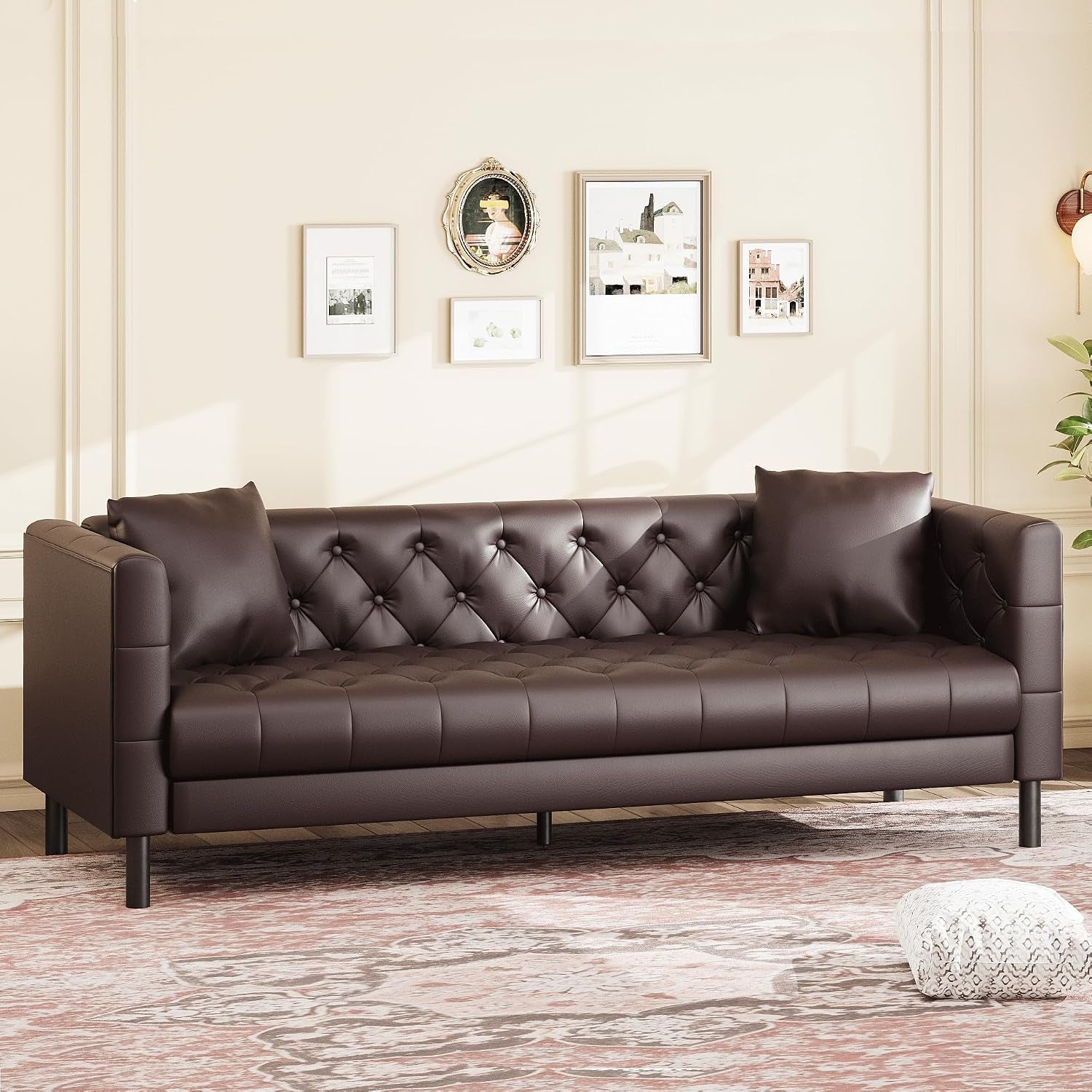 Front view of a dark brown tufted three-seat sofa with two pillows against a light wall with framed art.