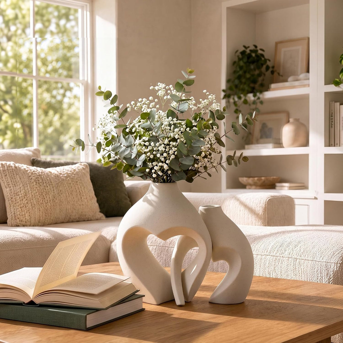 Large heart vase with eucalyptus stems and smaller heart vase on a bright living room coffee table beside an open book