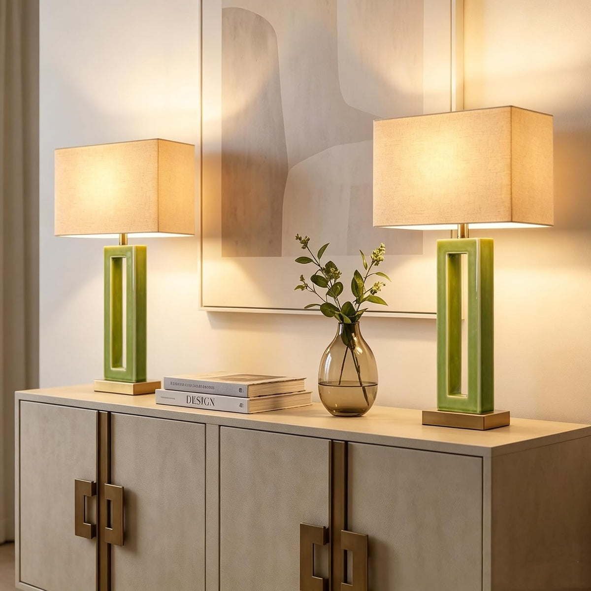Two lit green ceramic table lamps with beige rectangular shades on a console beside wall art and a small glass vase