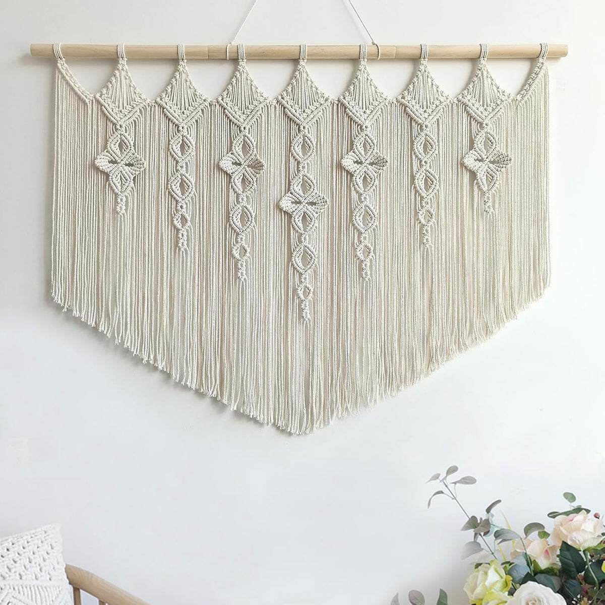 Macrame wall hanging on white wall with V-shaped fringe; lower corner shows a woven pillow and floral bouquet.