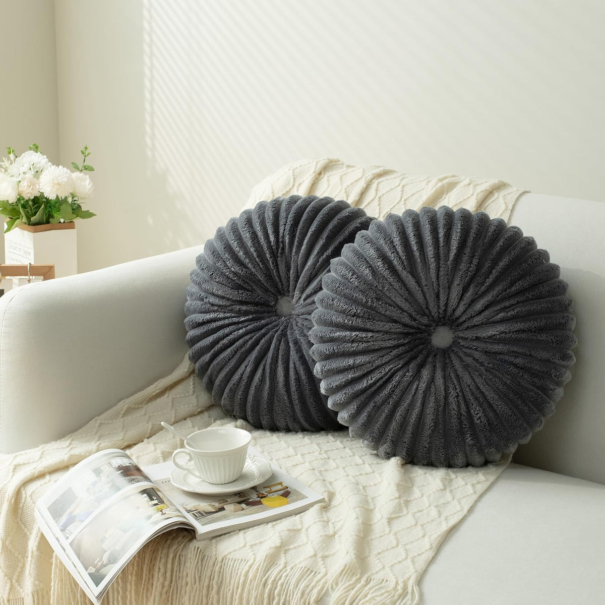 Two round ruched charcoal pillows on a sofa with a textured throw, teacup and flowers at the arm.
