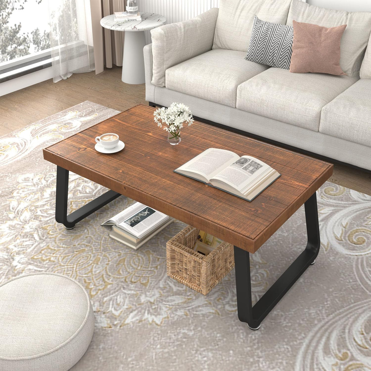 Top-down angle of wood coffee table with open book, small vase, woven basket and patterned rug beside a pouf.