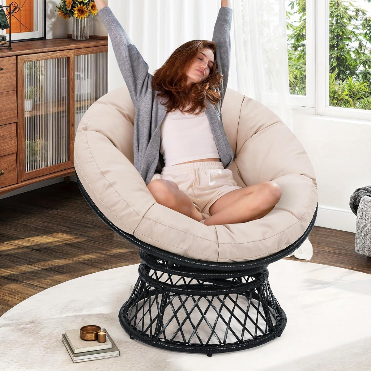 Woman stretching while seated in a beige cushioned papasan chair with black wicker base in a sunlit living room.