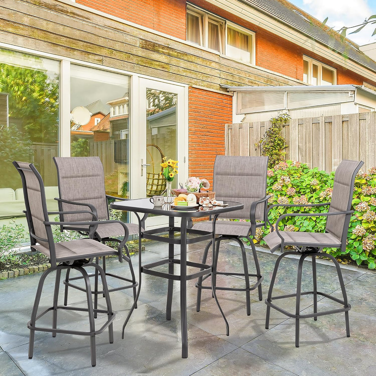 Four-piece patio bar set arranged on a backyard patio with brick house, plants and table laid for snacks.