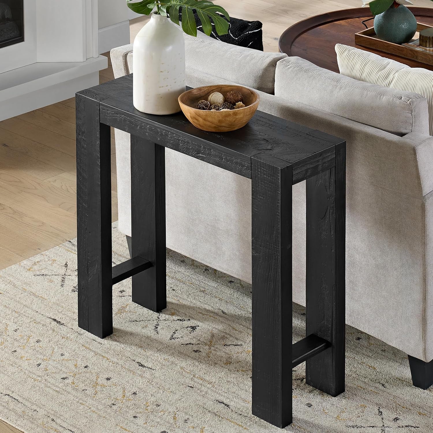 Narrow black console table behind a light gray sofa with a white vase and wooden bowl on top.