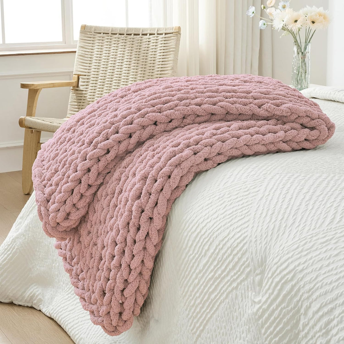 Folded dusty-pink chunky knit throw layered on the edge of a white bed with a woven chair nearby