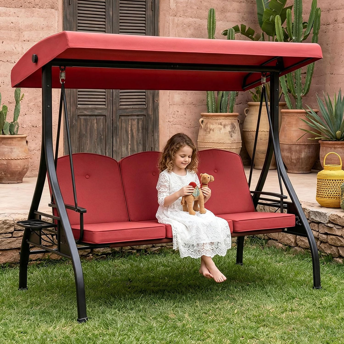 Red-cushioned porch swing on grass with a young girl in a white dress playing with a stuffed animal.